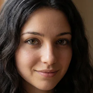 Alia Holes: a portrait. Long dark wavy hair, dark hazel eyes, a small smile, window light behind her.